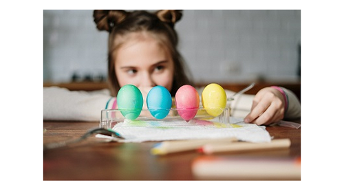 A girl sat at a table with 4 coloured eggs in front of her.