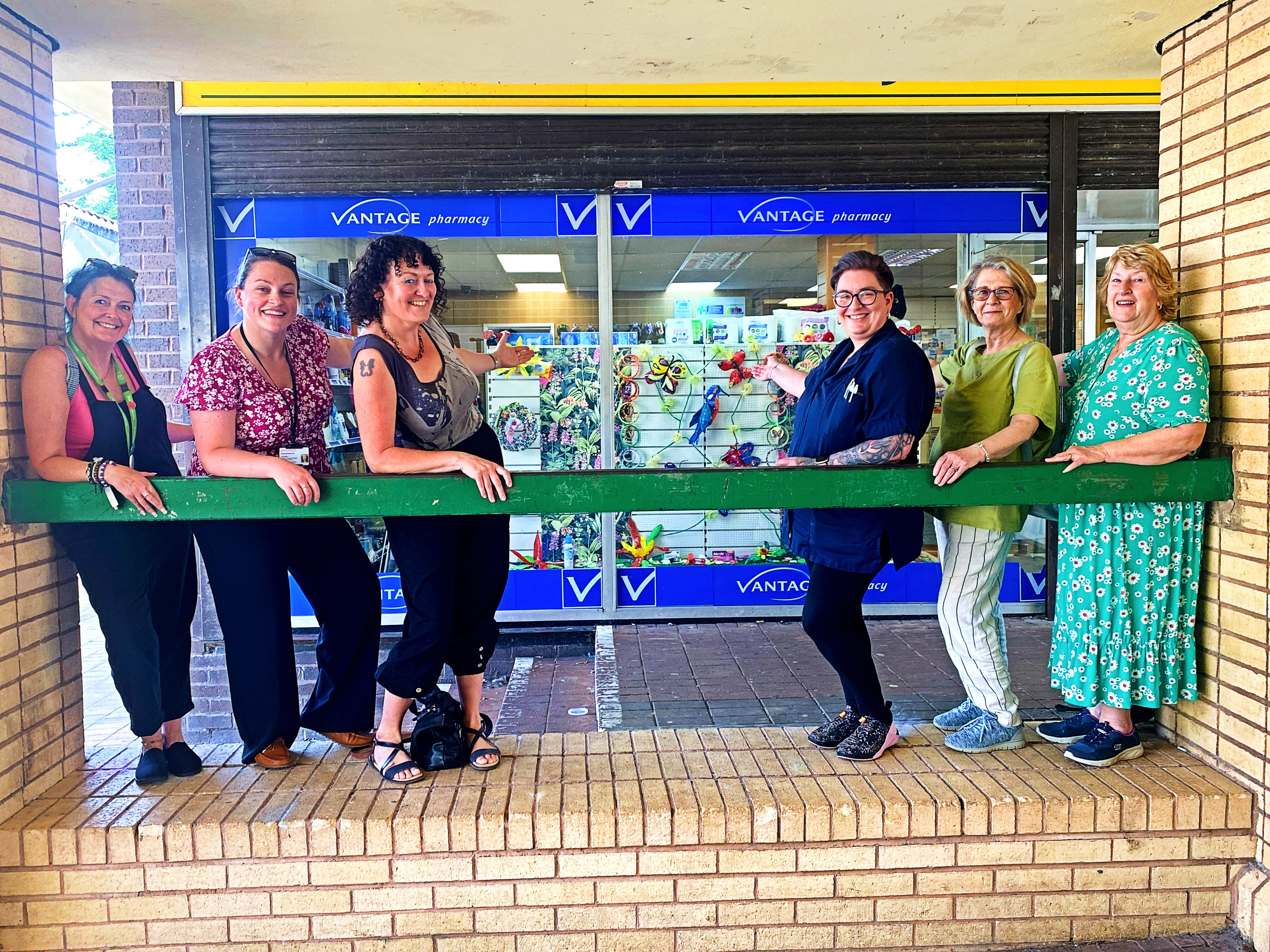 Six women stand in front of a pharmacy shop window that has been decorated with community art. 