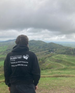 A photo of a young person stood onto top of a hill with their back to the camera. They are wearing a black hoodie with white text that says Worcestershire Children First fostering speak to us about fostering.