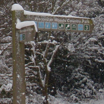 A photo of a wooden sign that reads Worcester Woods Country Park. The sign and trees in the background are covered with snow.