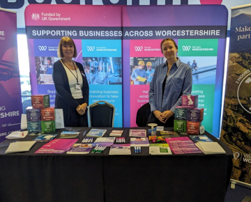 A photo of two people stood behind a table that has posters, leaflets and pens on it. Behind them are large posters with One Worcestershire and Visit Worcestershire branding.