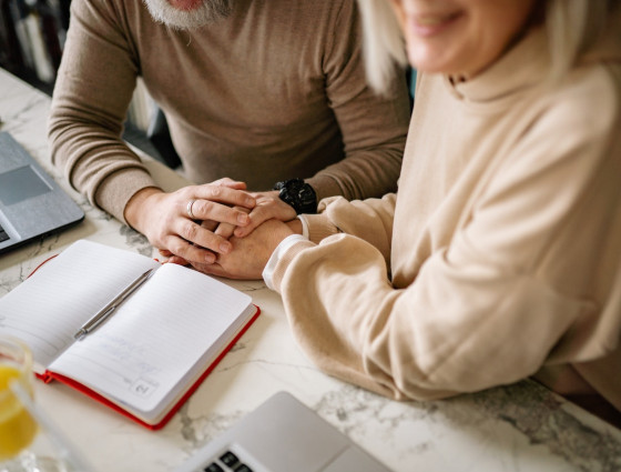 Close up of tow people holding hands sat at a table with a notepad on it