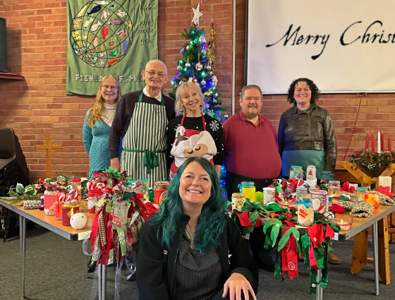 Five people stand in front of a Christmas tree with a table either side of them. Each table has handmade festive gifts on them. At the centre-front of the image a sixth person kneels facing the camera.