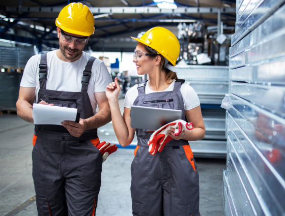 Male and Female factory workers in yellow hard hats holding clipboards walking and taking through factory.