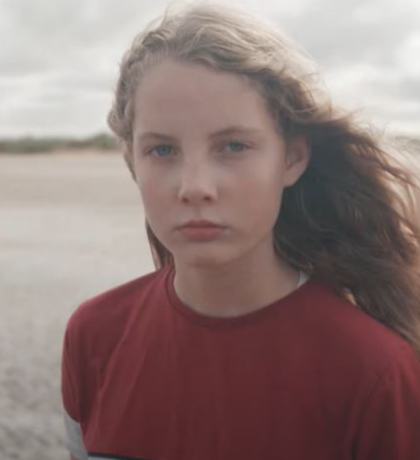 A still of a girl with long dark blonde hair in a red jumper standing on a beach.