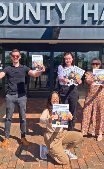 4 people stand outside the entrance to County Hall, each holding a copy of the INclusive Worcestershire magazine. There are 3 people stood at the rear (a man to the left and 2 women to his right) and a young woman kneels in the foreground facing the camera. 