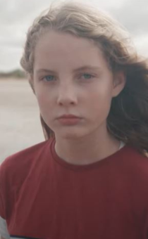 A still of a girl with long dark blonde hair in a red jumper standing on a beach.