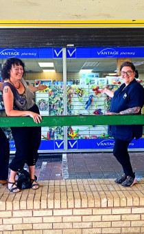 Six women stand in front of a pharmacy shop window that has been decorated with community art. 