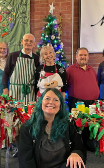 Five people stand in front of a Christmas tree with a table either side of them. Each table has handmade festive gifts on them. At the centre-front of the image a sixth person kneels facing the camera.