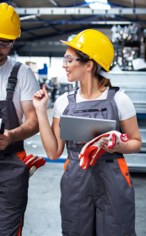 Male and Female factory workers in yellow hard hats holding clipboards walking and taking through factory.